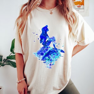 Puede incluir: Una camiseta de color crema con un dise&ntilde;o de acuarela de Ariel, la Sirenita, sentada en una roca con acuarelas azules y verdes.