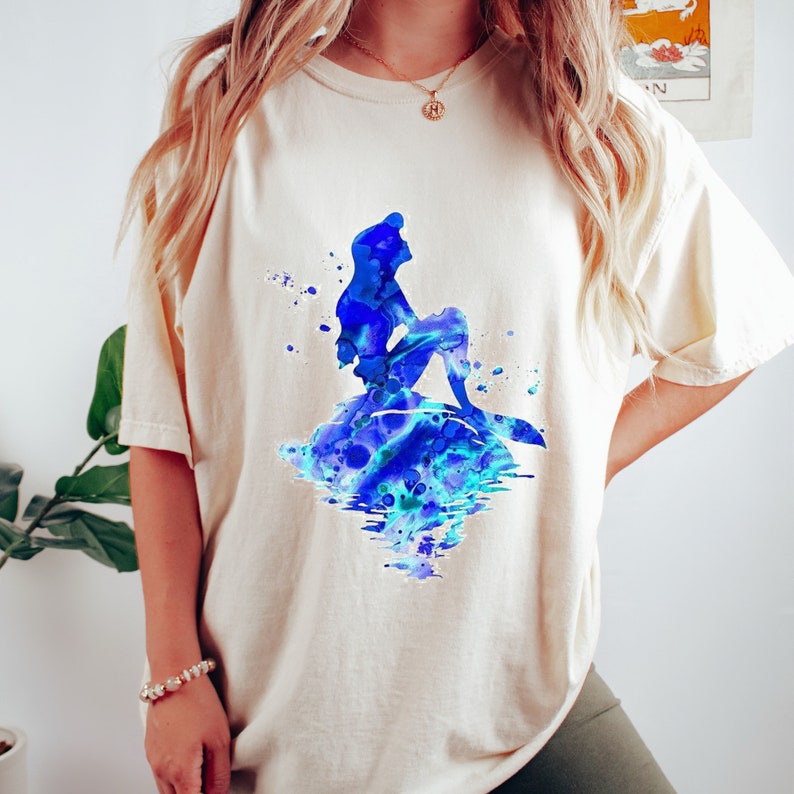 Puede incluir: Una camiseta de color crema con un dise&ntilde;o de acuarela de Ariel, la Sirenita, sentada en una roca con acuarelas azules y verdes.