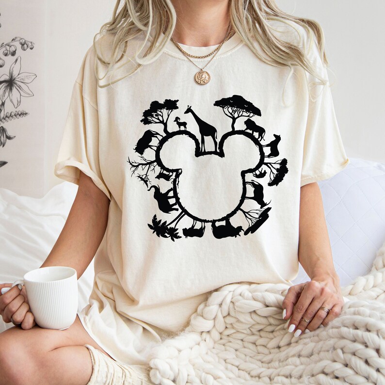 Puede incluir: Una camiseta blanca con una silueta negra de la cabeza de Mickey Mouse. La silueta est&aacute; formada por varios animales, entre ellos una jirafa, un le&oacute;n y una cebra, todos rodeados de &aacute;rboles.