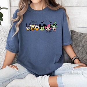 May include: A blue t-shirt featuring a graphic of Mickey Mouse and his friends dressed in Halloween costumes. The graphic includes Mickey Mouse, Minnie Mouse, Donald Duck, Goofy, Pluto, and a witch.