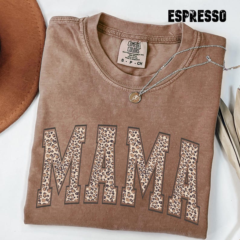 May include: A brown t-shirt with the word "MAMA" printed in a leopard print pattern. The shirt is made of a soft, comfortable material and has a relaxed fit.
