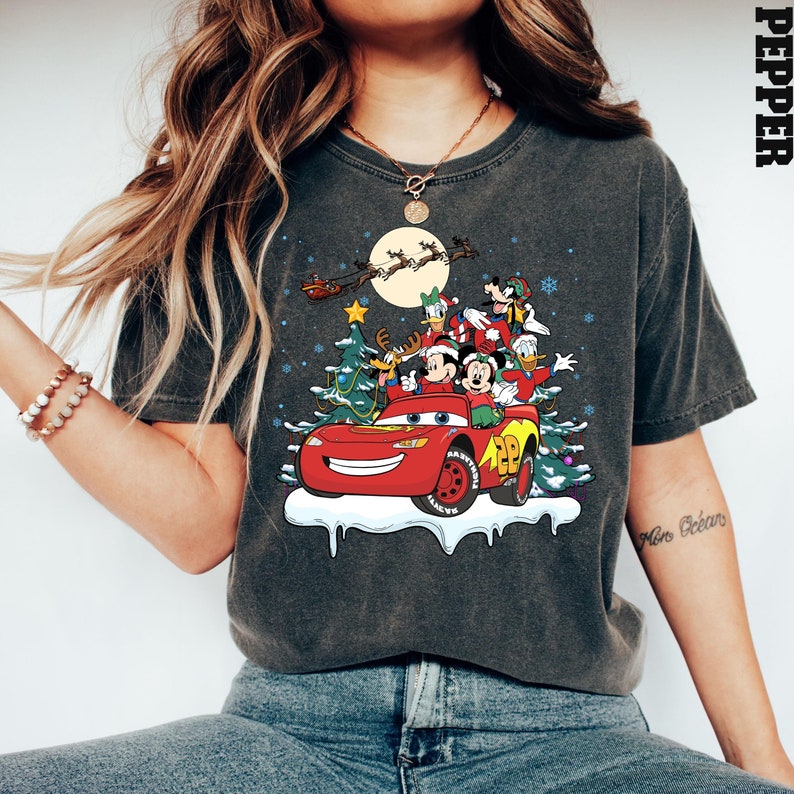 May include: A dark gray t-shirt with a Christmas-themed graphic featuring Mickey Mouse, Donald Duck, Goofy, and Pluto riding in a red Lightning McQueen car. The graphic also includes a Christmas tree, snowflakes, and a crescent moon.