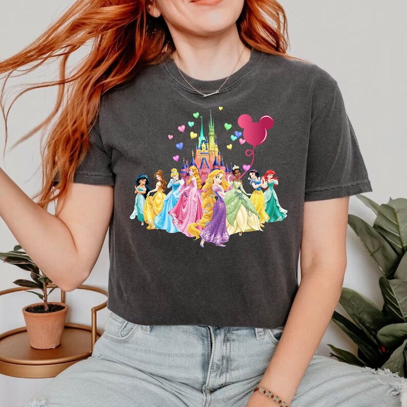 May include: A dark grey cropped t-shirt with a graphic of Disney princesses in front of Cinderella's castle with a pink Mickey Mouse balloon and hearts floating above.
