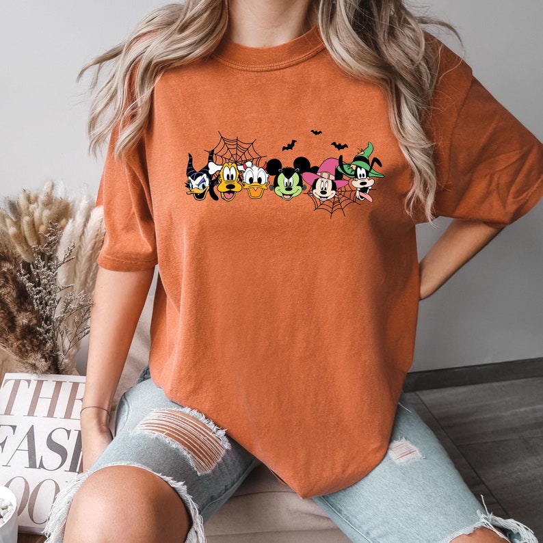 May include: Orange t-shirt with a Halloween-themed graphic featuring Mickey Mouse, Minnie Mouse, Donald Duck, Goofy, and other Disney characters. The characters are wearing costumes and are surrounded by bats and a spider web.