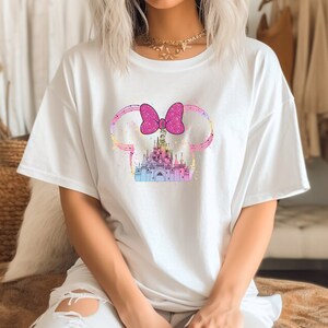 Puede incluir: Camiseta blanca con un lazo rosa y un gr&aacute;fico de castillo colorido. El castillo est&aacute; rodeado por un contorno rosa y azul que se asemeja a las orejas de Mickey Mouse.
