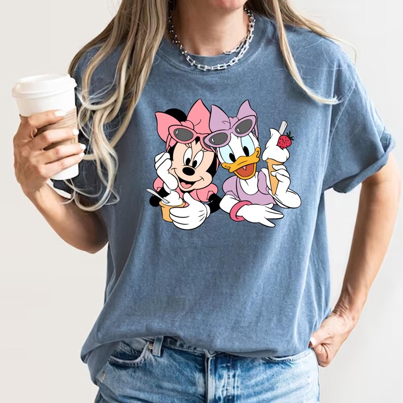 Puede incluir: Una camiseta azul con un gr&aacute;fico de Minnie Mouse y Daisy Duck con gafas de sol y sosteniendo conos de helado.