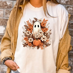 Op de afbeelding: Een wit T-shirt met een afbeelding van een witte geest omringd door oranje, bruine en witte bloemen en een kleine oranje pompoen.