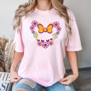 May include: Pink t-shirt with a floral design featuring a yellow bow and pink hearts. The design is in the shape of Mickey Mouse ears.
