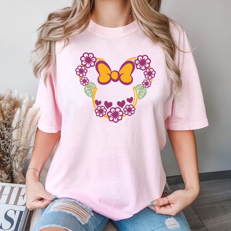May include: Pink t-shirt with a floral design featuring a yellow bow and pink hearts. The design is in the shape of Mickey Mouse ears.