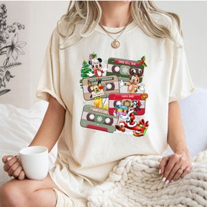 Puede incluir: Una camiseta blanca con un dise&ntilde;o navide&ntilde;o con Mickey Mouse y otros personajes de Disney. El dise&ntilde;o incluye cintas de cassette con t&iacute;tulos de canciones como "Jingle Bell Rock", "Frosty the Snowman" y "Santa Baby".
