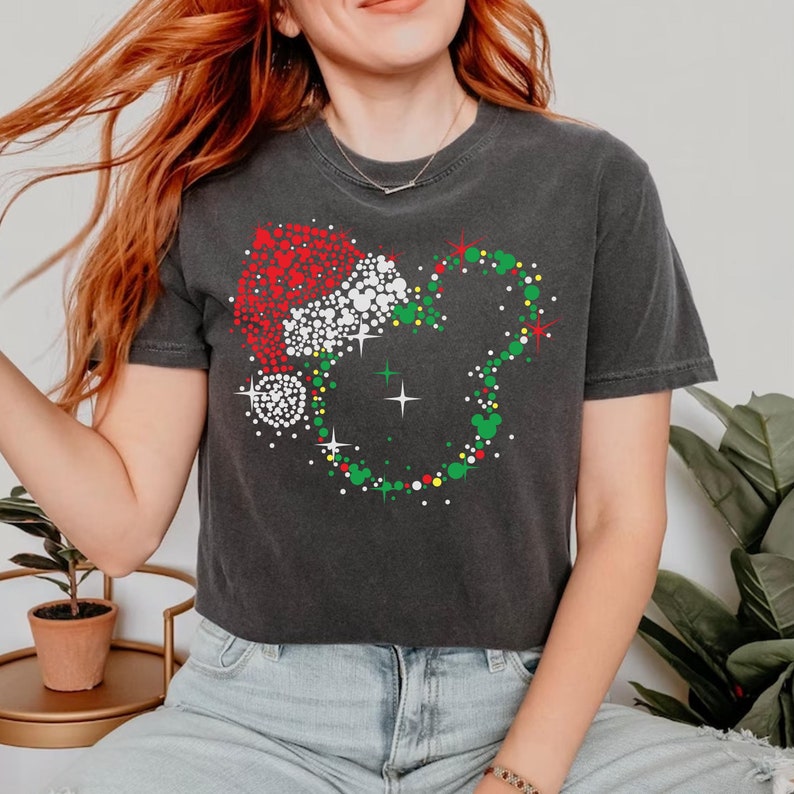 Puede incluir: Camiseta gris oscuro con un dise&ntilde;o navide&ntilde;o. El gr&aacute;fico incluye un gorro de Pap&aacute; Noel y una corona con forma de Mickey Mouse, ambos compuestos por puntos de colores. El dise&ntilde;o incorpora elementos rojos, blancos, verdes y amarillos, creando un tema festivo.