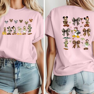 May include: Two pink t-shirts with a cartoon character design. The front shirt features a line of Disney characters with balloons. The back shirt features a variety of Disney characters with bows and a jungle theme.