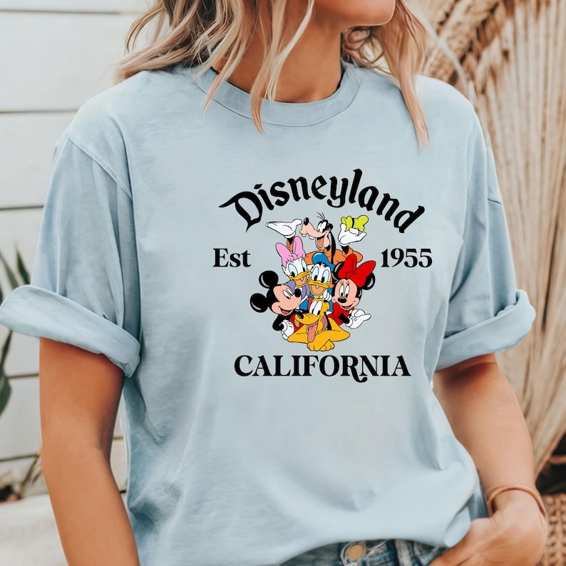 Puede incluir: Camiseta azul claro con un gr&aacute;fico negro de Mickey Mouse y sus amigos. El gr&aacute;fico incluye el texto "Disneyland Est 1955 California".