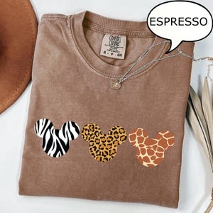Pu&ograve; includere: T-shirt color espresso con tre disegni a forma di testa di Topolino con motivi animalier: zebra, leopardo e giraffa. La maglietta ha girocollo e maniche corte. Visibile una collana d'argento con un piccolo ciondolo.
