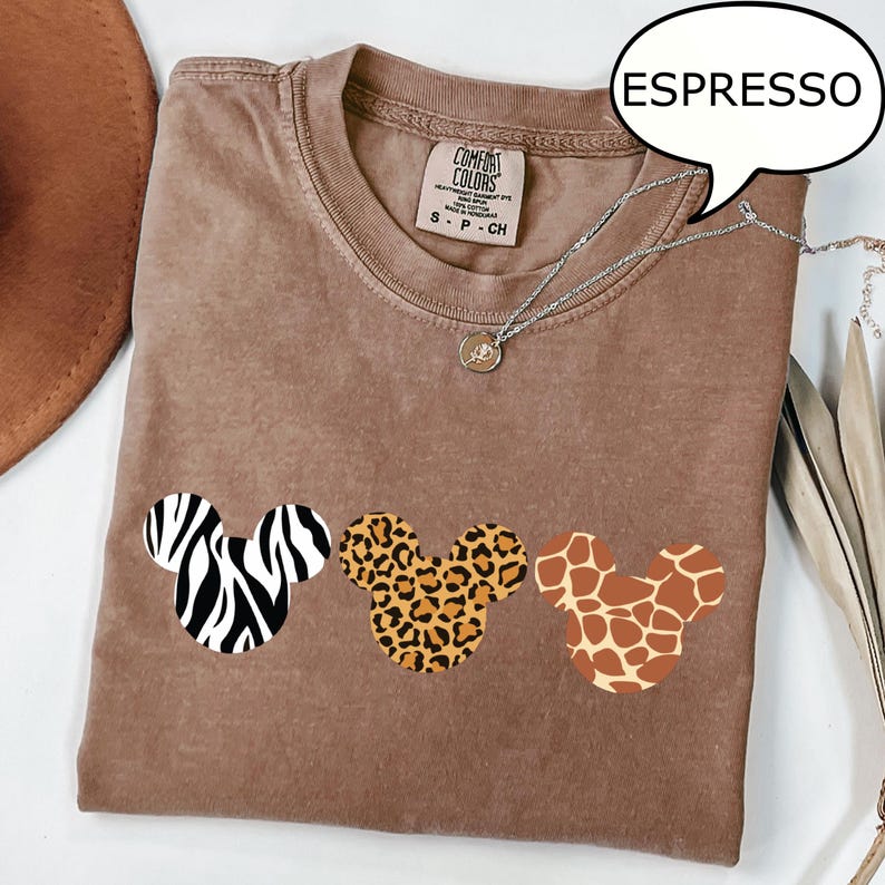 Pu&ograve; includere: T-shirt color espresso con tre disegni a forma di testa di Topolino con motivi animalier: zebra, leopardo e giraffa. La maglietta ha girocollo e maniche corte. Visibile una collana d'argento con un piccolo ciondolo.