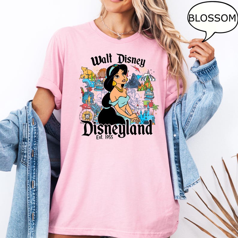 May include: Pale pink t-shirt with a graphic of Princess Jasmine from Disney's Aladdin, and the text "Walt Disney Disneyland Est. 1955". The design includes various park icons in a colourful illustration.