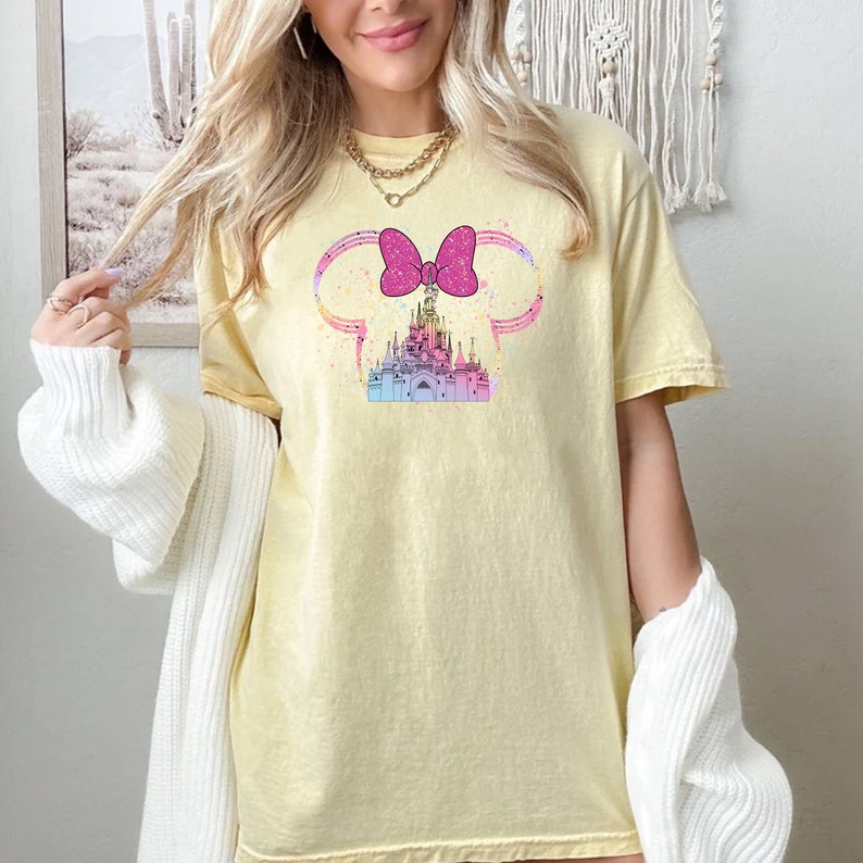 Puede incluir: Una camiseta amarilla con un lazo rosa y un gr&aacute;fico de castillo en colores pastel. El castillo est&aacute; dentro de un par de orejas de Mickey Mouse.