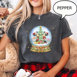 May include: Dark gray t-shirt featuring a snow globe design with Mickey and Minnie Mouse in Christmas attire. The snow globe includes a Christmas tree, gifts, and candy canes. The word "PEPPER" is in a speech bubble.