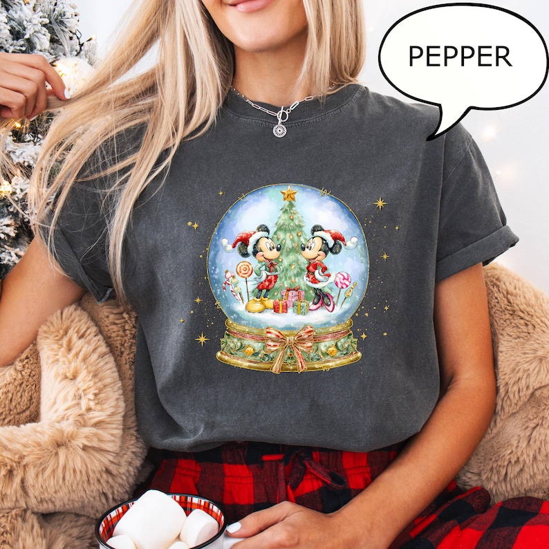 May include: Dark gray t-shirt featuring a snow globe design with Mickey and Minnie Mouse in Christmas attire. The snow globe includes a Christmas tree, gifts, and candy canes. The word "PEPPER" is in a speech bubble.
