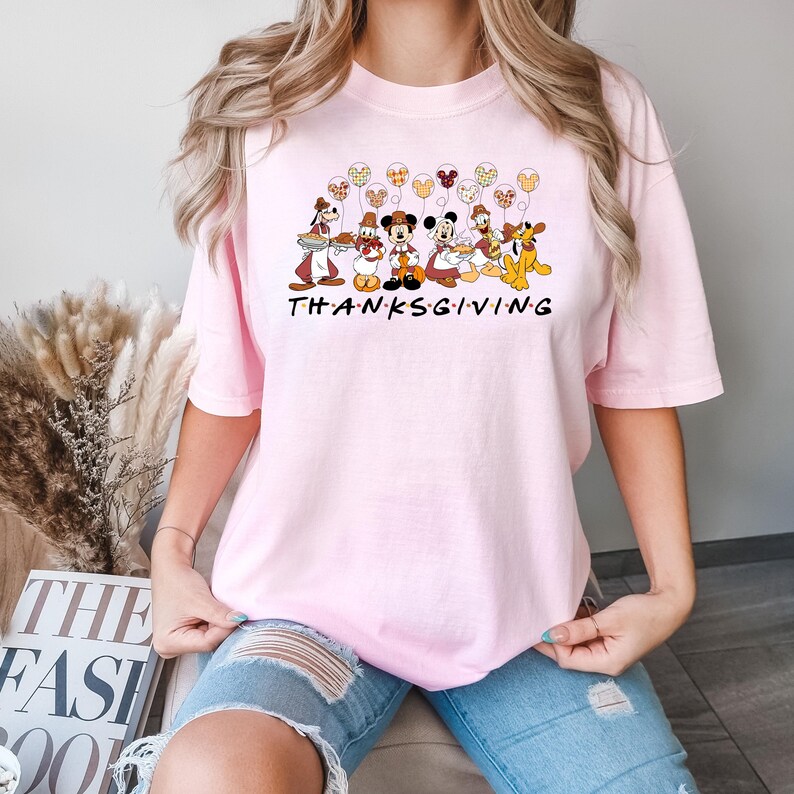 May include: Pink t-shirt with a graphic of Disney characters holding Thanksgiving themed items and the word "THANKSGIVING" printed below.