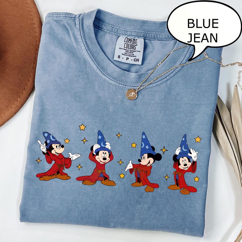 May include: Blue jean colored t-shirt featuring four Mickey Mouse characters dressed as wizards. Each character wears a blue wizard hat and a red robe. The shirt has a Comfort Colors tag and a gold necklace.