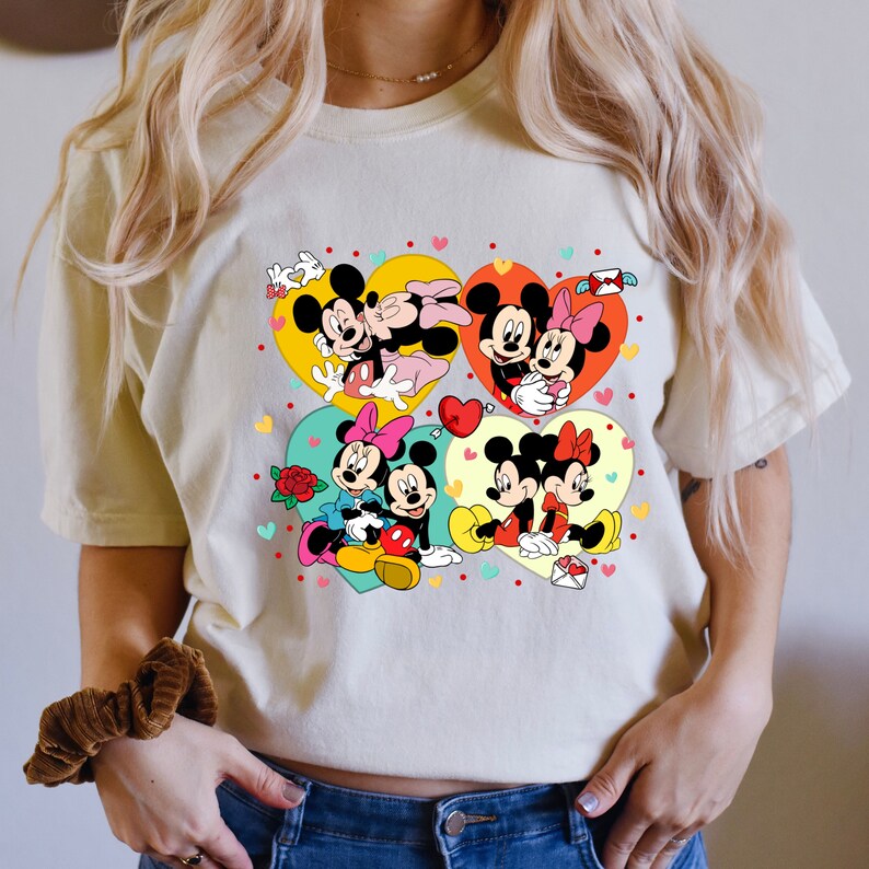 Puede incluir: Una camiseta blanca con un estampado gr&aacute;fico colorido de Mickey Mouse y Minnie Mouse en varias poses. El gr&aacute;fico presenta corazones y otros s&iacute;mbolos rom&aacute;nticos.