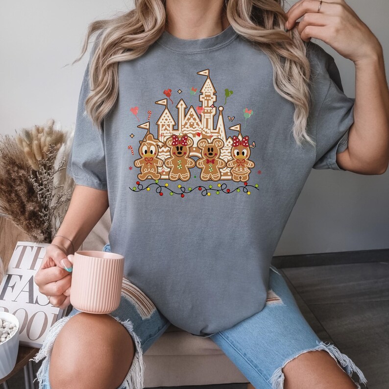 May include: Gray t-shirt featuring a gingerbread castle and four gingerbread people. The gingerbread figures have red bows and are decorated with icing. The design includes colorful lights and hearts, creating a festive holiday theme.