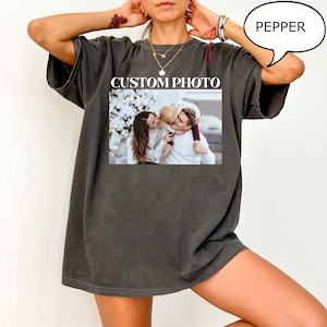 May include: Dark gray t-shirt with a printed photo. The photo shows a family of three. The text "CUSTOM PHOTO" is printed above the photo. The word "PEPPER" is in a speech bubble.
