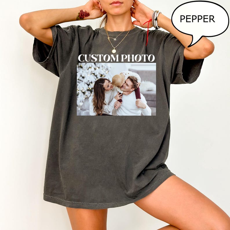 May include: Dark gray t-shirt with a printed photo. The photo shows a family of three. The text "CUSTOM PHOTO" is printed above the photo. The word "PEPPER" is in a speech bubble.