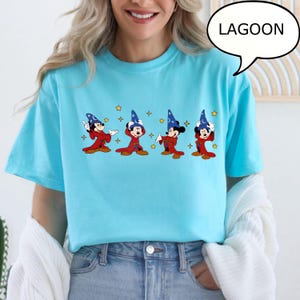 May include: A light blue t-shirt featuring a cartoon design of Mickey Mouse in a wizard costume. The design includes four Mickey Mouse figures in red robes and blue wizard hats, with stars. The word "LAGOON" is in a speech bubble.