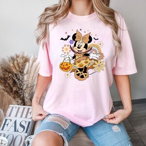 Puede incluir: Camiseta rosa claro con un personaje de dibujos animados de Halloween sosteniendo una calabaza. El dise&ntilde;o incluye un arco&iacute;ris, flores, murci&eacute;lagos y estrellas. La camiseta es de corte holgado.