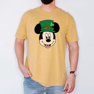 Puede incluir: Camiseta de color amarillo mostaza con la cara de Mickey Mouse de dibujos animados con un sombrero de duende verde con un tr&eacute;bol. El gr&aacute;fico est&aacute; centrado en la camiseta. La camiseta tiene mangas cortas y cuello redondo.