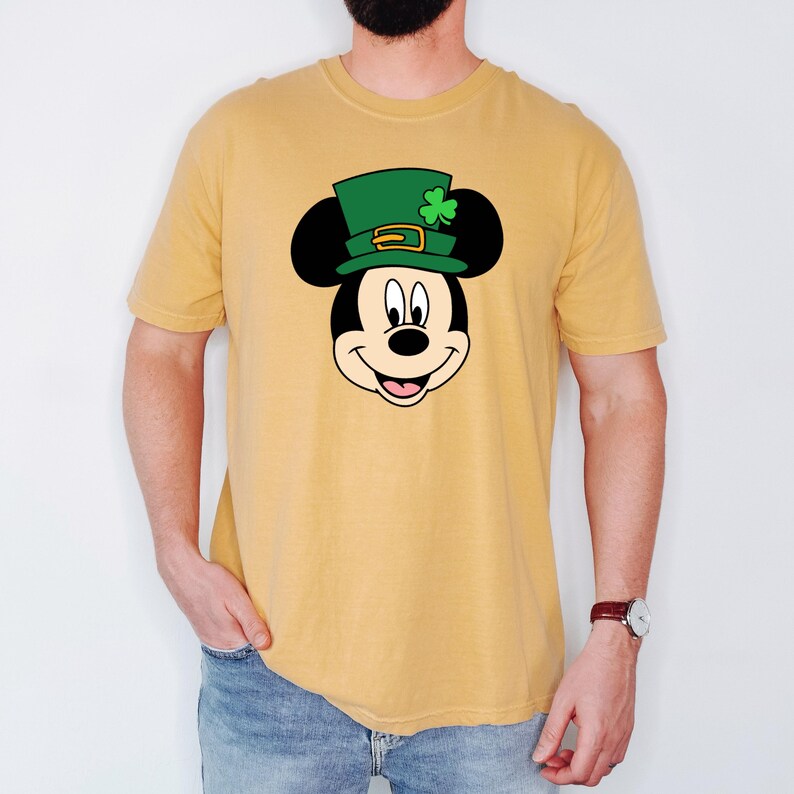 Puede incluir: Camiseta de color amarillo mostaza con la cara de Mickey Mouse de dibujos animados con un sombrero de duende verde con un tr&eacute;bol. El gr&aacute;fico est&aacute; centrado en la camiseta. La camiseta tiene mangas cortas y cuello redondo.