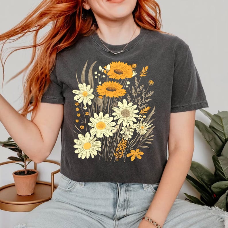 May include: Dark gray t-shirt featuring a floral design with yellow sunflowers and white daisies. The graphic includes various other wildflowers and foliage. The shirt has short sleeves and a crew neck.