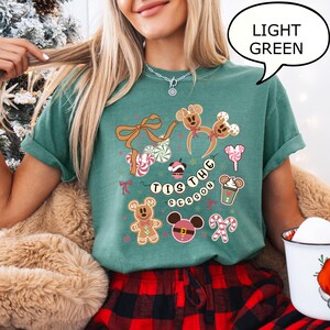 May include: Light green t-shirt with a festive design featuring gingerbread men, candy canes, and the phrase "Tis the Season." The shirt has a crew neck and short sleeves, perfect for the holiday season.