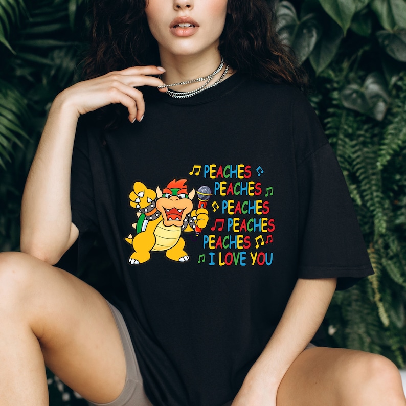 May include: Black t-shirt featuring a cartoon Bowser character holding a microphone. The shirt has the text "PEACHES" repeated with musical notes and "I LOVE YOU" in colorful letters. The design is centered on the front of the shirt.