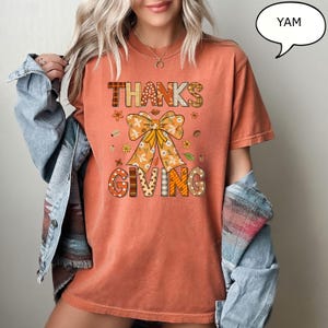 May include: An orange t-shirt with the words "THANKS GIVING" in a decorative font. The design includes a large bow and floral accents, perfect for Thanksgiving celebrations. The shirt is a comfortable, casual style.