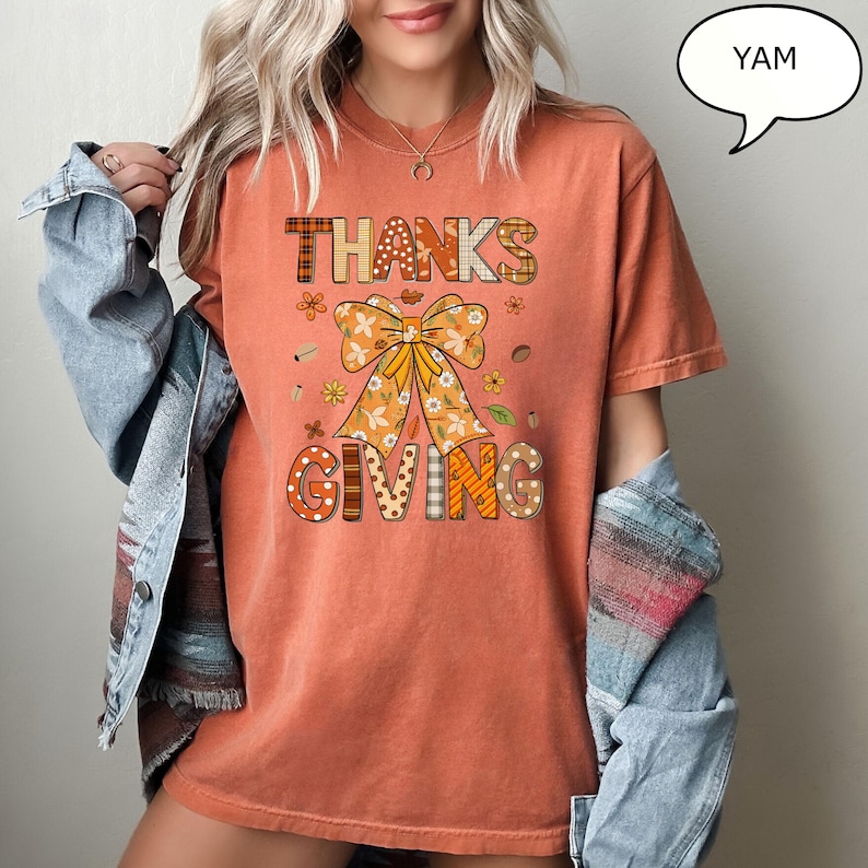 May include: An orange t-shirt with the words "THANKS GIVING" in a decorative font. The design includes a large bow and floral accents, perfect for Thanksgiving celebrations. The shirt is a comfortable, casual style.