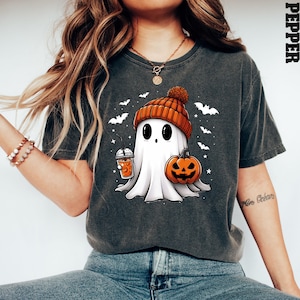 May include: A dark grey t-shirt featuring a cartoon ghost wearing an orange and brown hat, holding a pumpkin and a cup of orange liquid. The ghost is surrounded by bats and stars. The text "Mom Goals" is on the right sleeve of the shirt.