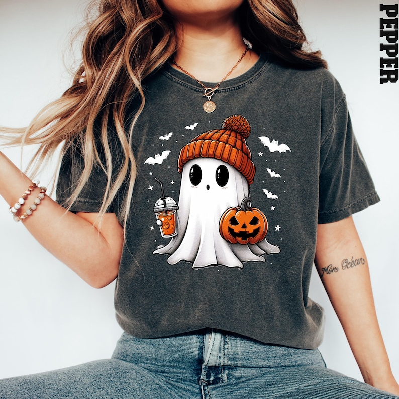 May include: A dark grey t-shirt featuring a cartoon ghost wearing an orange and brown hat, holding a pumpkin and a cup of orange liquid. The ghost is surrounded by bats and stars. The text "Mom Goals" is on the right sleeve of the shirt.