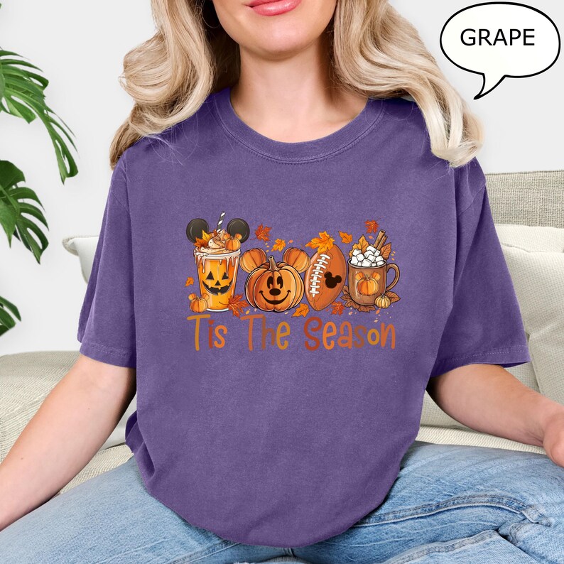 May include: A purple t-shirt with a fall-themed graphic. The graphic includes a pumpkin, a football, and a mug of hot chocolate, all with Mickey Mouse ears. The text "T's The Season" is printed below the graphic.