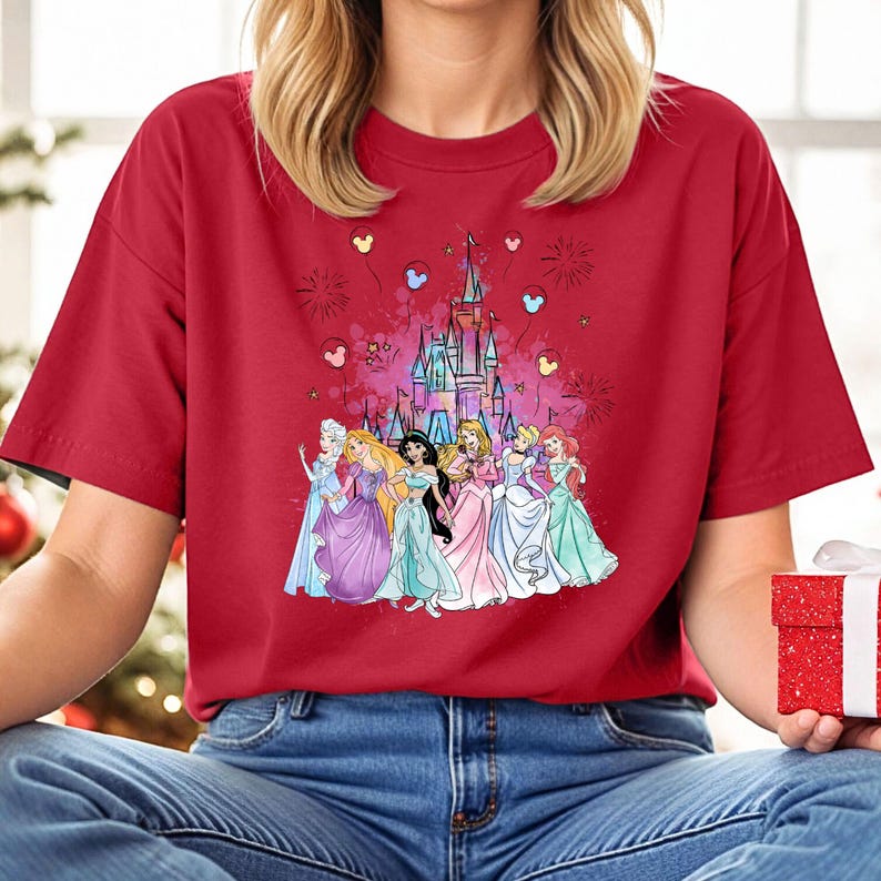 Puede incluir: Camiseta roja con un gr&aacute;fico del castillo de Disney y siete princesas Disney delante de &eacute;l. Hay globos y fuegos artificiales en el fondo.