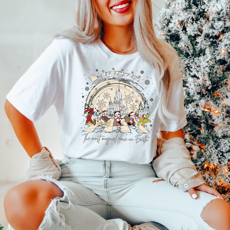 Puede incluir: Camiseta blanca con un gr&aacute;fico de Mickey Mouse y sus amigos caminando por una calle con un fondo navide&ntilde;o. El texto "The most magical place on Earth" est&aacute; impreso debajo del gr&aacute;fico.