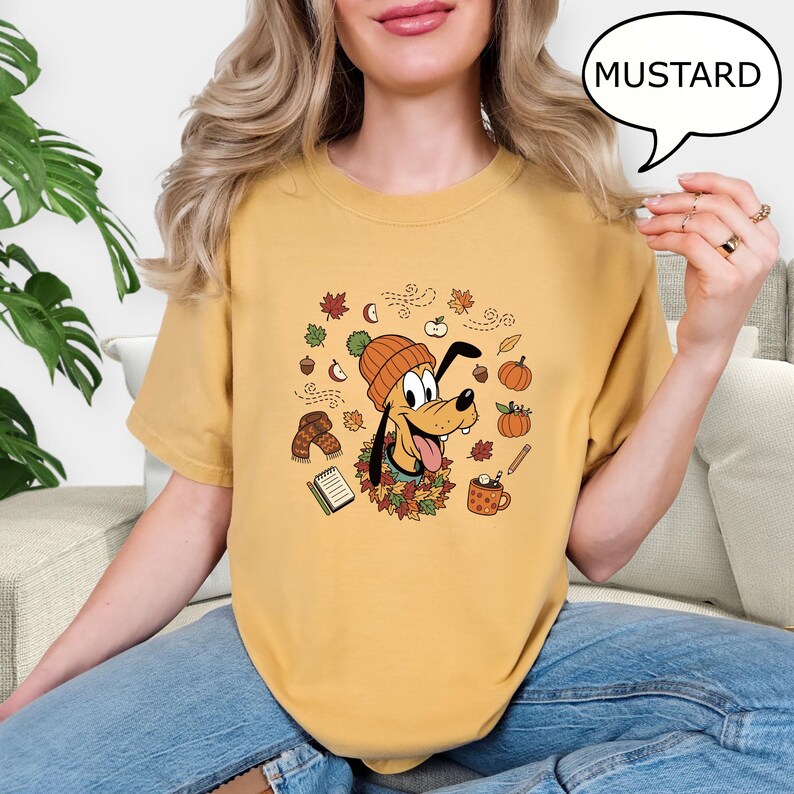 May include: Mustard-colored t-shirt featuring a cartoon dog wearing a knit cap and surrounded by fall-themed graphics. The dog is surrounded by autumn leaves, pumpkins, and a mug of hot cocoa. The word "MUSTARD" is in a speech bubble.