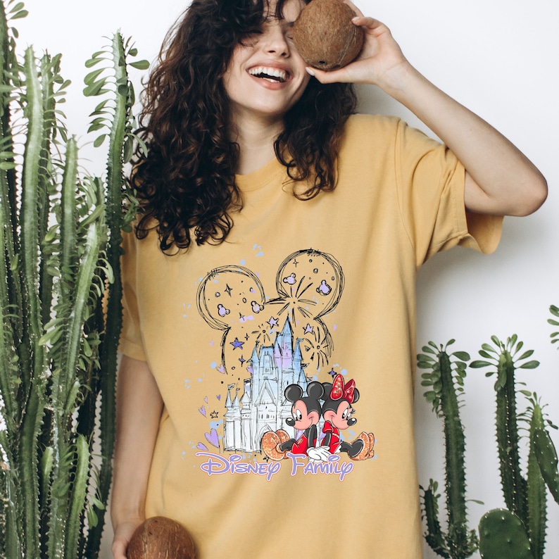 May include: A yellow t-shirt featuring a cartoon illustration of Mickey Mouse and Minnie Mouse in front of Cinderella's Castle. The illustration is surrounded by fireworks and stars. The text "Disney Family" is printed below the illustration.