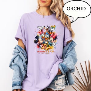 May include: Orchid-colored t-shirt featuring a colorful graphic of Mickey & Co. characters, including Mickey Mouse, Minnie Mouse, Donald Duck, and Goofy. The text "Mickey & Co. EST 1928" is printed below the characters. The word "ORCHID" is in a speech bubble.