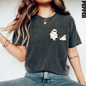 May include: A dark grey t-shirt featuring a white ghost wearing a yellow floral pattern and a smaller white ghost dog on a lead. The text "Pepper" is in the top right corner of the image.
