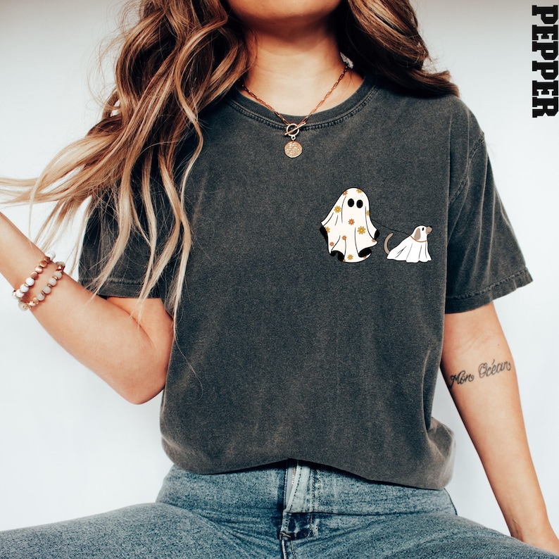 May include: A dark grey t-shirt featuring a white ghost wearing a yellow floral pattern and a smaller white ghost dog on a lead. The text "Pepper" is in the top right corner of the image.