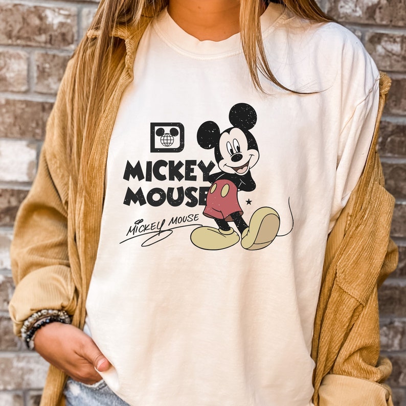 May include: White t-shirt with a vintage Mickey Mouse graphic. The graphic features Mickey Mouse in a red and black outfit with a yellow star on his shorts. The text "Mickey Mouse" is printed in black letters below the graphic.