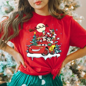 May include: Red t-shirt with a Christmas scene featuring Mickey Mouse, Donald Duck, Goofy, and Pluto riding in a red car with a Christmas tree on top. The car is driving through a snowy scene with a full moon in the background.